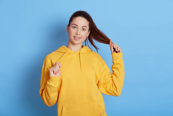 Portrait of smiling playful young woman with dark hair and ponytail, wears yellow sweatshirt, isolated over blue background, looks at camera with charming smile, being in good mood. - Фото, зображення