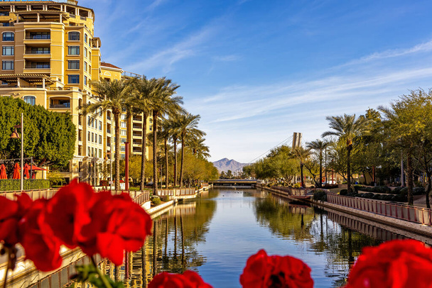 Tagsüber läuft ein Kanal durch das Wohn- und Einkaufsviertel der Altstadt von Scottsdale, Arizona, USA. Umrahmt von defokussierten roten Blumen. - Foto, Bild