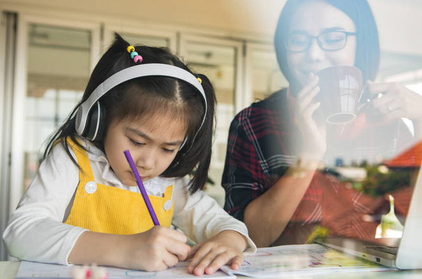 Madre asiática sosteniendo la taza de café y y mira a la pequeña hija que hace la tarea a su lado, Ella utiliza lápiz para escribir en el libro. - Foto, Imagen