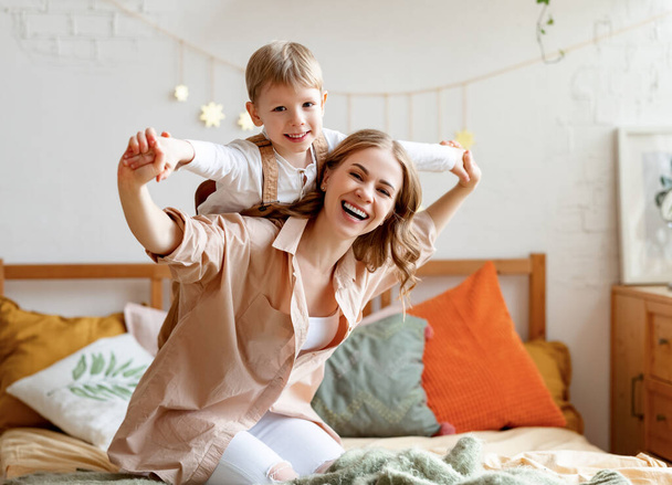 Podekscytowana matka śmiejąc się i niosąc wesoły chłopiec na plecach podczas klęcząc na łóżku w przytulnej sypialni w weekend w domu - Zdjęcie, obraz