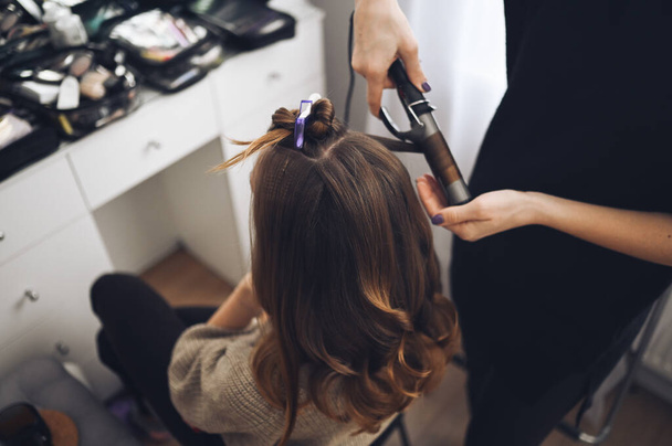 Stylista włosów przygotowuje kobieta sprawia, że loki fryzurę z lokówki. Długie jasnobrązowe naturalne włosy - Zdjęcie, obraz