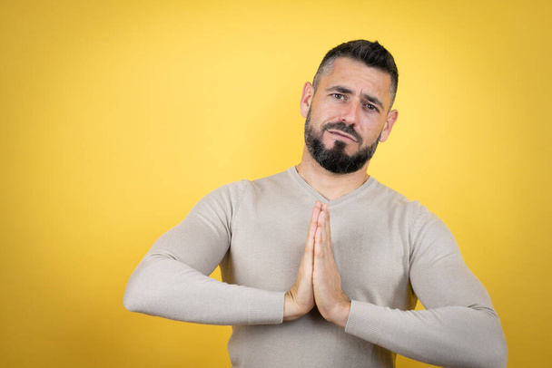 Красавчик с бородой в свитере на жёлтом фоне просит и молится руками вместе с выражением надежды на лице очень эмоциональным и взволнованным - Фото, изображение