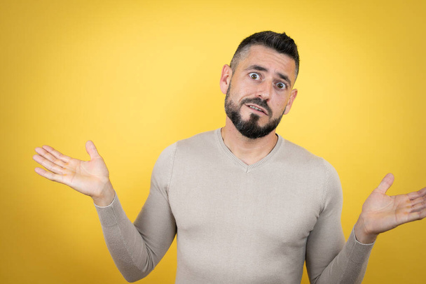 Handsome man with beard wearing sweater over yellow background clueless and confused expression with arms and hands raised - Photo, Image