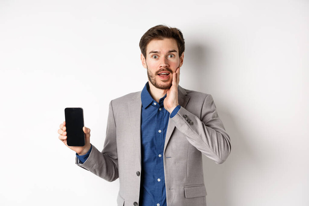 Homme d'affaires excité en costume montrant écran vide smartphone et haletant étonné, démontrer l'application, debout sur fond blanc - Photo, image