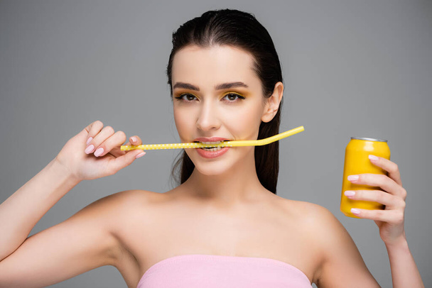 brunette young woman holding yellow can with soft drink and biting straw isolated on grey  - Photo, Image