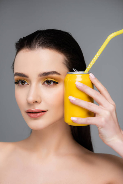 brunette jeune femme tenant boîte jaune avec boisson gazeuse avec paille près du visage isolé sur gris  - Photo, image