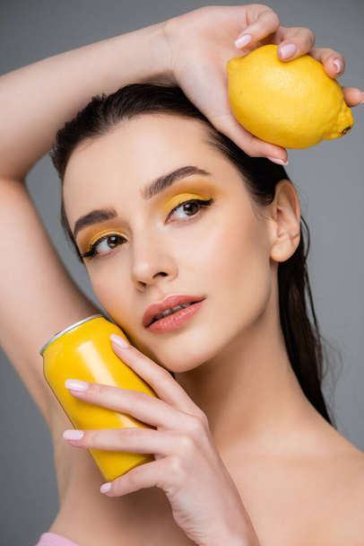 mujer joven sosteniendo lata amarilla con refresco y limón maduro aislado en gris  - Foto, imagen
