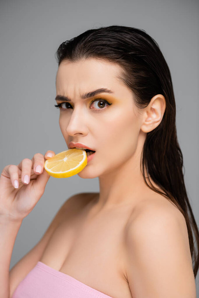 surprised young woman with bare shoulders biting sliced lemon isolated on grey  - Foto, Imagen