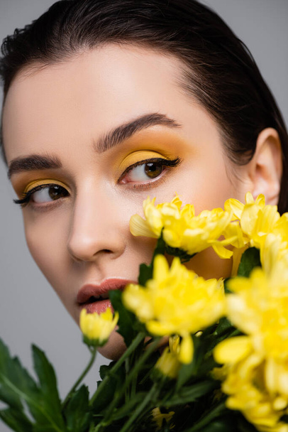 młoda brunetka kobieta z makijażem w pobliżu kwitnące żółte kwiaty na rozmytym pierwszym planie odizolowany na szarym - Zdjęcie, obraz
