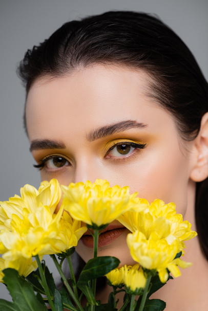 young woman with yellow eyeshadows covering face with blooming flowers isolated on grey - Photo, Image