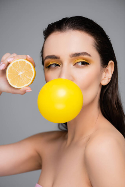mujer joven con el maquillaje que sopla la goma de mascar amarilla y la celebración de la mitad de limón aislado en gris - Foto, imagen