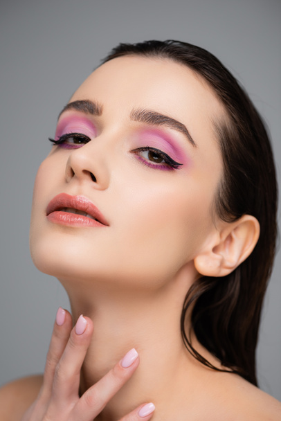 young woman with pink eye shadows and bare shoulders looking at camera isolated on grey  - Foto, Imagem