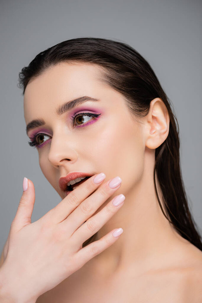 νεαρή γυναίκα με ροζ σκιές ματιών και γυμνούς ώμους αγγίζοντας τα χείλη απομονώνονται σε γκρι  - Φωτογραφία, εικόνα