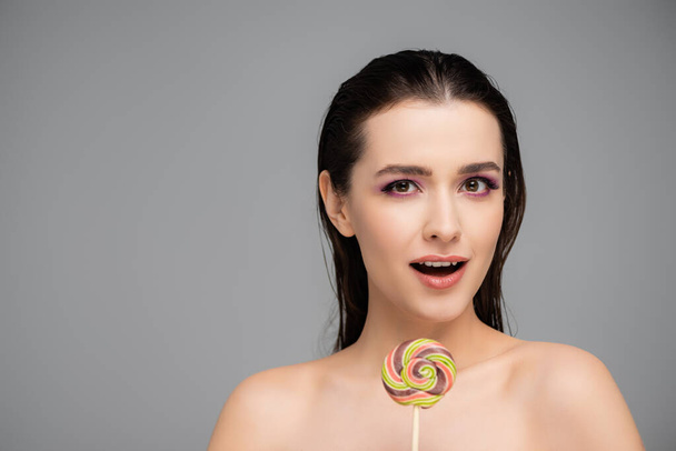 surprised young woman with pink eye shadows holding lollipop isolated on grey  - Photo, Image
