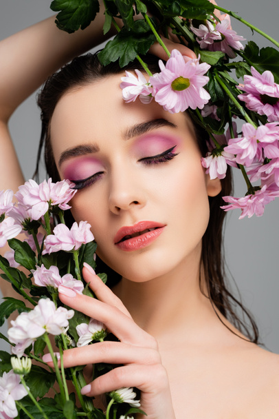 młoda kobieta z zamkniętymi oczami i różowymi cieniami do powiek trzymająca kwiaty odizolowane na szaro  - Zdjęcie, obraz