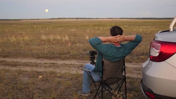 A man traveler in car stopped at a camping in evening and takes pictures with a camera next to car. Freelance photographer working outdoors, beautiful moments for photos and videos. Driver is resting - Photo, Image