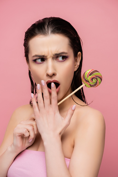 schockierte junge Frau bedeckt Mund und hält Lutscher isoliert auf rosa  - Foto, Bild