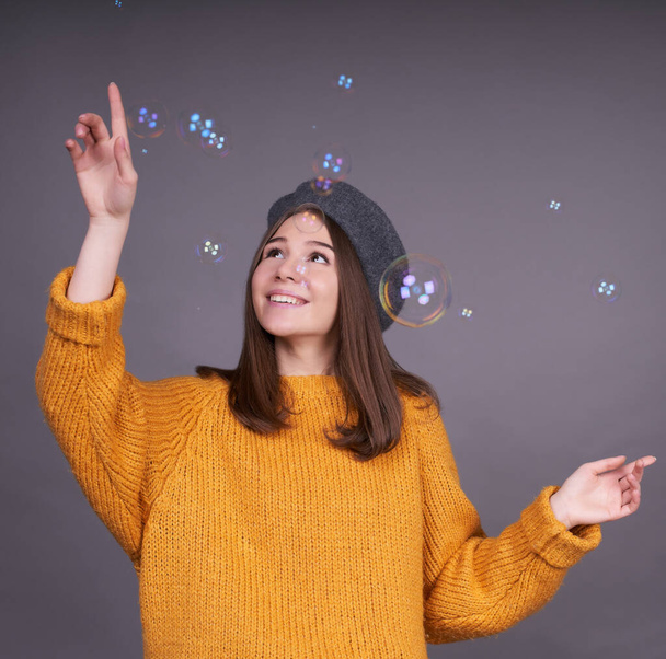 Young attractive Caucasian beautiful woman in a yellow sweater and a gray beret, stands on a gray background, catches soap bubbles with her hands. - Fotoğraf, Görsel