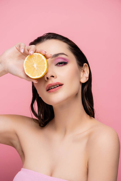 Genç bir kadın, çıplak omuzlarıyla gözlerini yarım limonla kapatıyor ve pembe renkli kameraya bakıyor. - Fotoğraf, Görsel