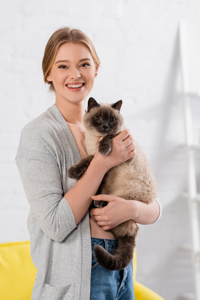 Mujer feliz mirando a la cámara mientras sostiene gato siamés en casa  - Foto, Imagen