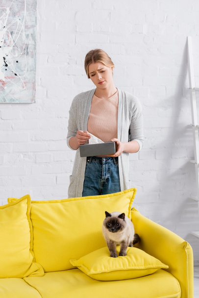 Mujer triste sosteniendo servilleta y caja cerca del gato siamés en el sofá  - Foto, Imagen