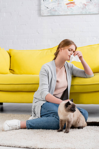 Young woman with napkin feeling allergy reaction near siamese cat on carpet  - Φωτογραφία, εικόνα