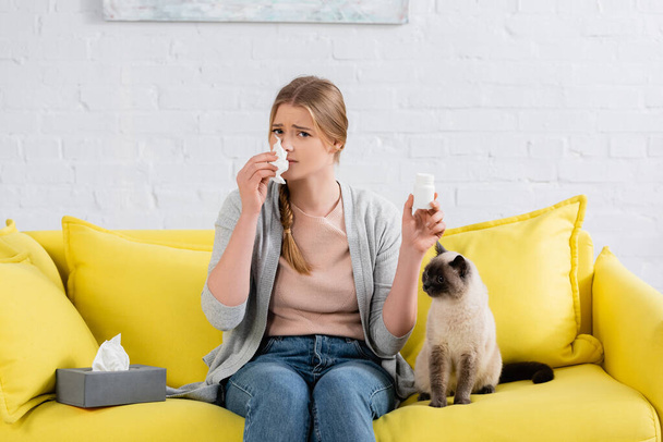 Vrouw met allergie houden servet en pillen in de buurt van siamese kat op de bank  - Foto, afbeelding