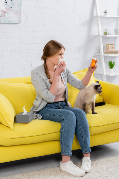 Mujer con alergia mirando frasco con pastillas cerca de gato en sofá amarillo en casa  - Foto, Imagen