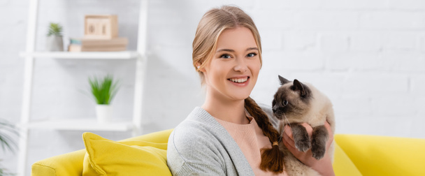 Cheerful woman smiling at camera while holding siamese cat at home, banner  - Fotoğraf, Görsel
