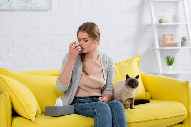 Frau mit Allergie mit Inhalator in der Nähe pelziger siamesischer Katze auf Sofa  - Foto, Bild