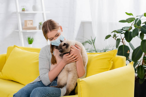 Junge Frau mit Allergie-Reaktion trägt medizinische Maske und hält pelzige Katze auf gelbem Sofa  - Foto, Bild