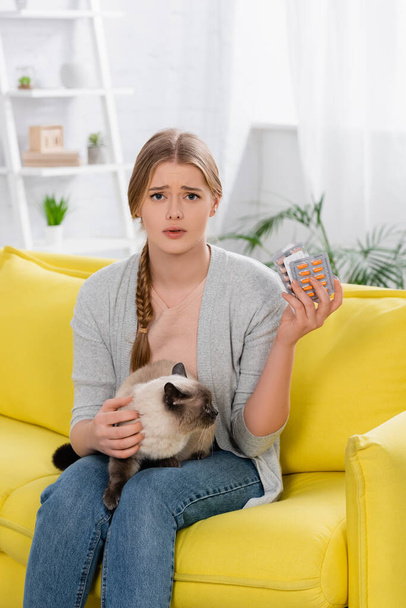 Mujer molesta con alergia sosteniendo ampollas con pastillas y gato siamés en el sofá  - Foto, imagen