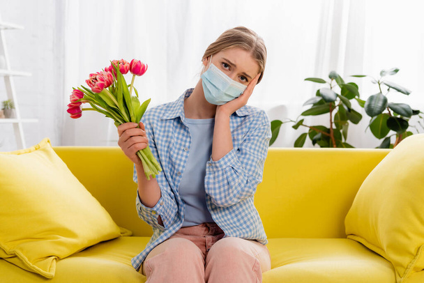 Θλιβερή γυναίκα με ιατρική μάσκα που κρατά τουλίπες κατά τη διάρκεια αλλεργίας  - Φωτογραφία, εικόνα