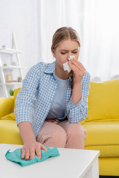 Woman with allergy having snuffle while cleaning table on blurred foreground  - Photo, Image