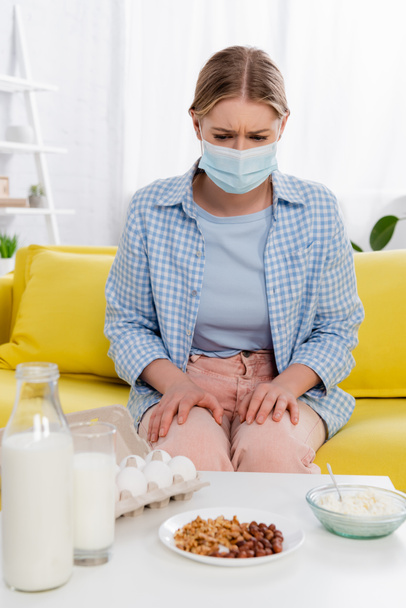 Woman with allergy wearing medical mask near milk, cottage cheese and eggs on blurred foreground  - Photo, Image