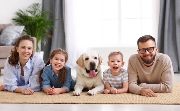 Usmívající se matka a otec s malými dětmi a rozkošný čistokrevný labradorský retrívr pes ležící na podlaze a dívající se na kameru, zatímco tráví čas spolu doma - Fotografie, Obrázek
