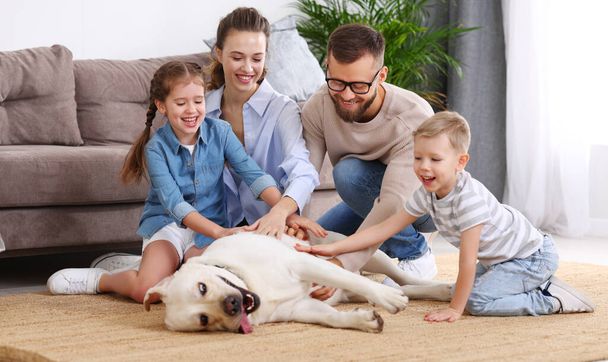 Padres felices y niños pequeños acariciando adorable perro cansado acostado en la alfombra mientras disfrutan de fin de semana juntos en casa - Foto, imagen