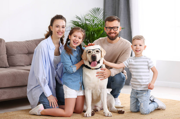 Onnelliset vanhemmat ja lapset kokoontuvat ihastuttavan puhdasrotuisen labradorinnoutajan koiran ympärille hauskoissa silmälaseissa ja katsomalla kameraa nauttien viikonlopusta yhdessä kotona. - Valokuva, kuva