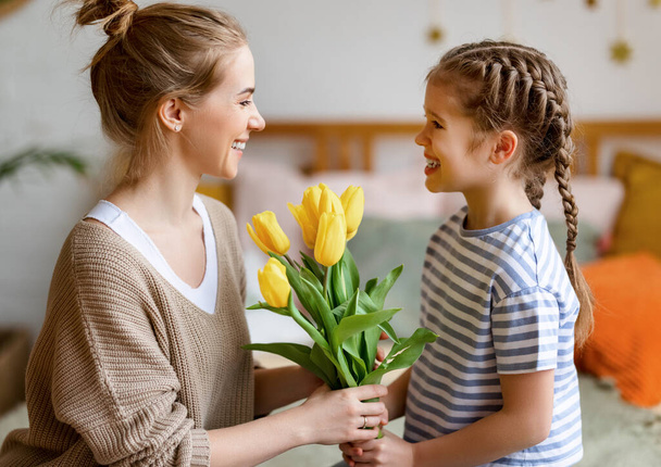Vista lateral de la hija alegre felicita a su madre en las vacaciones del día de la madre y le da un ramo de flores, mientras que felicita en vacaciones en casa - Foto, imagen