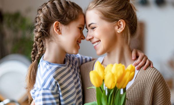 Оптимистичная семья: мама с букетом белых тюльпанов, улыбающихся и трогательных лба со счастливой дочерью во время праздника празднования Дня матери дома - Фото, изображение