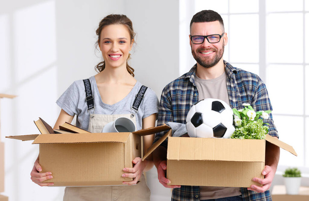 Feliz joven hombre y mujer sonriendo y mirando a la cámara mientras lleva cajas de cartón con pertenencias durante la reubicación en un nuevo apartamento - Foto, Imagen