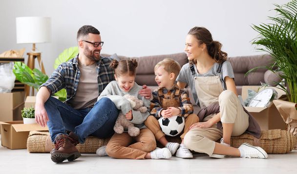 Γοητευμένη οικογένεια: γονείς και χαριτωμένα παιδιά συγκεντρώνονται στο πάτωμα στο σαλόνι με κουτιά unpacked σε νέο διαμέρισμα και κοιτάζοντας την κάμερα - Φωτογραφία, εικόνα