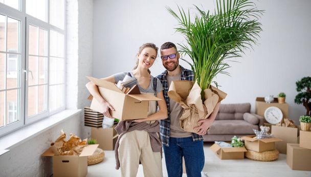Зміст молода пара стоїть з картонними коробками і вазоном в новій квартирі і насолоджується переїздом, дивлячись на камеру
 - Фото, зображення