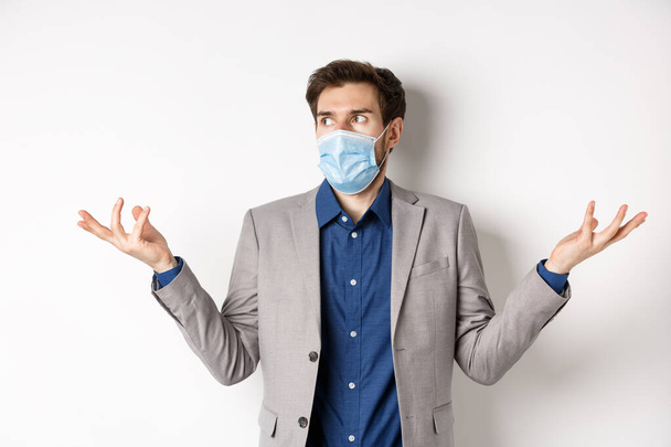 Confused businessman in medical mask shrugging and looking aside, know nothing, cant understand something, standing on white background - Photo, Image