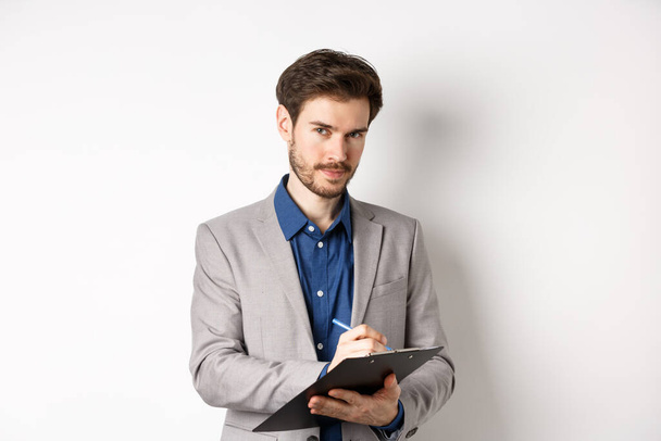Homme d'affaires prospère en costume gris prenant des notes, travaillant dans les vêtements de bureau, tenant le presse-papiers et regardant la caméra, fond blanc - Photo, image