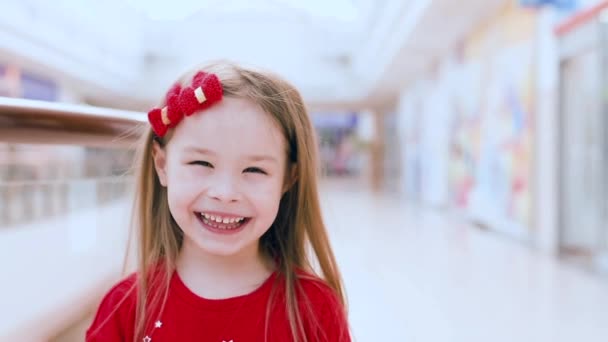 Küçük kız kameraya bakıp gülüyor. Bir kız alışveriş merkezinde duruyor ve gülümsüyor. - Video, Çekim