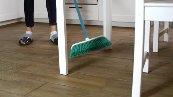 台所の床を掃除するために、現代のブロムスティックを使用して自宅で家事を行うカジュアルな服装の若い女性。スリッパとほうきで若い女性の足 - 映像、動画