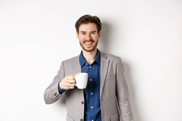 Красивий бізнесмен у костюмі, який п'є каву або чай з офісного горнятка, посміхаючись на камеру, стоїть на білому тлі
 - Фото, зображення