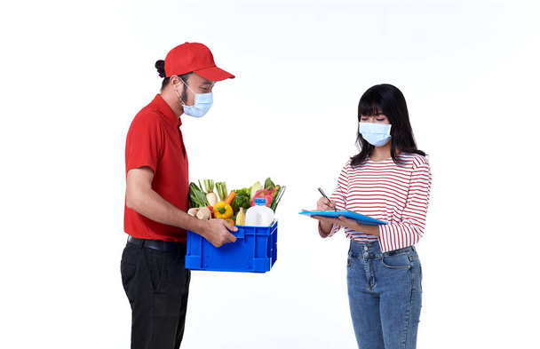 Ασιάτης ντελιβεράς φορώντας μάσκα προσώπου σε κόκκινη στολή με παντοπωλείο κουτί τροφίμων, φρούτων, λαχανικών και ποτών δώσει σε γυναίκα πελάτη απομονώνονται σε λευκό φόντο. κοινωνική αποστασιοποίηση και νέα κανονική έννοια. - Φωτογραφία, εικόνα