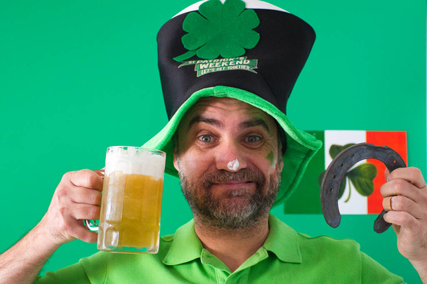 Человек с кружкой свежего, пенного пива со шляпой и подковою в руке и зеленым фоном празднует День Святого Патриция - Фото, изображение